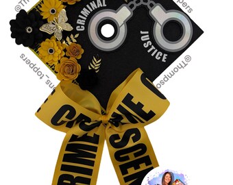 Pode incluir: Tampo de formatura preto decorado com flores de papel amarelas e pretas, borboletas e detalhes dourados. O tampo apresenta as palavras "CRIMINAL JUSTICE" e um desenho de algemas. Uma fita amarela com o texto "CRIME SCENE" está amarrada ao tampo.