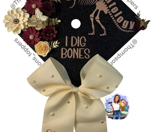 Pode incluir: Tampão de formatura preto decorado com um esqueleto de dinossauro, a palavra "Paleontology" e a frase "I DIG BONES". O tampão é adornado com flores de papel creme e bordô e um laço creme com pérolas. O laço diz "Turma de 2025".