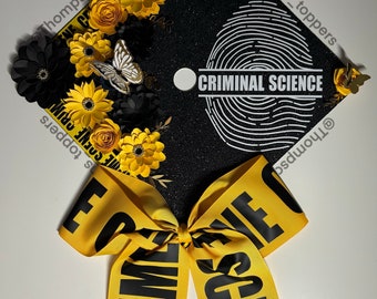 Pode incluir: Um chapéu de formatura preto decorado com flores de papel amarelas e pretas, uma borboleta prateada e uma fita amarela com as palavras "CRIME SCENE". O chapéu apresenta o texto "CRIMINAL SCIENCE" e um desenho de impressão digital.