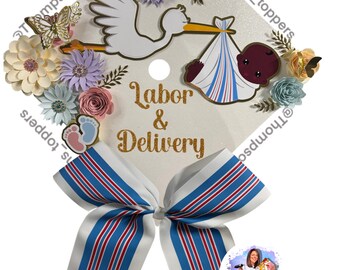 Pode incluir: Um chapéu de formatura branco decorado com uma cegonha carregando um bebê, rodeado por flores de papel em cores pastel. As palavras "Labor & Delivery" estão em glitter dourado. Um laço com listras azuis e vermelhas completa o design.