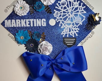 Pode incluir: Um chapéu de formatura azul decorado com flores de papel azuis, brancas e douradas, um laço azul com detalhes dourados e um gráfico de lâmpada com a palavra "MARKETING" e vários ícones de marketing. O chapéu é coberto com glitter azul.