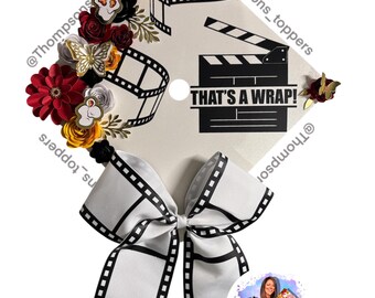 Pode incluir: Um chapéu de formatura decorado com um tema de filme. O chapéu apresenta um laço branco com um design de tira de filme, um gráfico de claquete e o texto "THAT'S A WRAP!". É adornado com flores vermelhas, amarelas e douradas e uma borboleta.