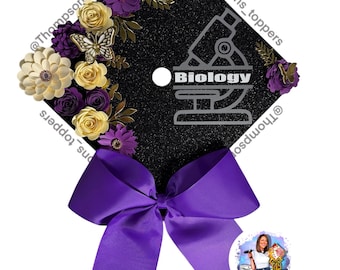 Pode incluir: Um chapéu de formatura preto decorado com flores de papel em creme, dourado e roxo, e um laço roxo. O chapéu apresenta a palavra "Biologia" e um gráfico de microscópio. O chapéu é adornado com uma borboleta.