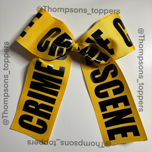 May include: A yellow ribbon bow with the words "CRIME SCENE" printed in bold black letters. The bow is centered and has two long tails. The text is clear and easy to read.