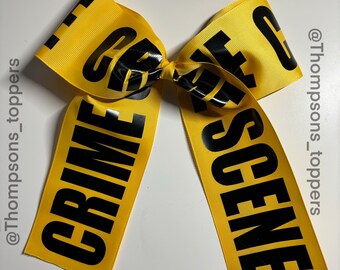 Pode incluir: Um laço de fita amarelo com as palavras "CRIME SCENE" impressas em letras pretas em negrito. O laço é amarrado no centro e tem caudas longas e fluidas. O texto está centrado na fita.