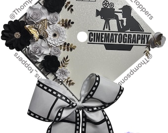 Puede incluir: Un gorro de graduación plateado decorado con flores blancas y negras, una silueta negra de una persona sosteniendo una cámara y un lazo de cinta negra y blanca con diseño de rollo de película. El gorro también presenta el texto "2025" y "CINEMATOGRAFÍA".
