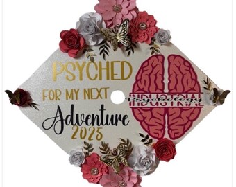 Puede incluir: Un gorro de graduación decorado con las palabras "PSYCHED FOR MY NEXT Adventure 2025" en oro y negro. El gorro presenta un gráfico de cerebro y está adornado con flores de papel y mariposas.