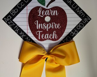 Pode incluir: Decoração de chapéu de formatura com uma maçã vermelha brilhante que diz "Learn Inspire Teach". O chapéu tem um laço amarelo com um desenho de lápis. As bordas são pretas com letras brancas.
