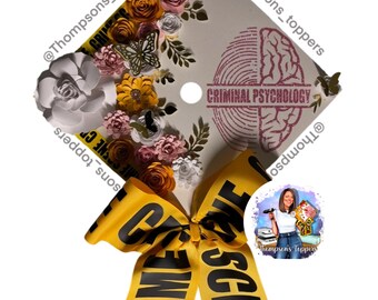 Pode incluir: Um chapéu de formatura decorado com um arranjo floral de flores de papel em branco, amarelo, rosa e laranja. O chapéu apresenta um gráfico de cérebro e as palavras "CRIMINAL PSYCHOLOGY". Uma fita amarela com o texto "CRIME SCENE" está amarrada em um laço.