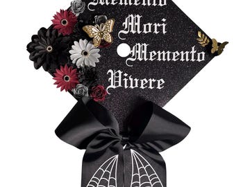 Puede incluir: Gorra de graduación negra con purpurina decorada con flores rojas, negras y blancas. La gorra presenta la frase "Memento Mori Memento Vivere" en escritura blanca, con un lazo negro y diseño de telaraña.