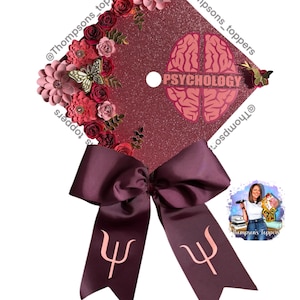 May include: Burgundy glitter graduation cap decorated with paper flowers, butterflies, and a bow. The cap features a brain graphic and the word "PSYCHOLOGY". A ribbon with the psychology symbol hangs from the bow.