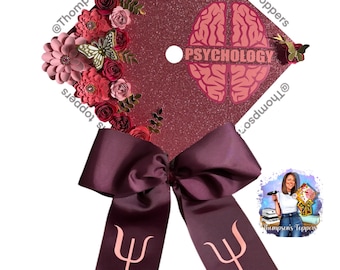 Pode incluir: Tampo de formatura com glitter bordô decorado com flores de papel, borboletas e um laço. O tampo apresenta um gráfico de cérebro e a palavra "PSYCHOLOGY". Uma fita com o símbolo da psicologia pende do laço.