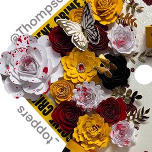 Puede incluir: Un arreglo decorativo con flores de papel en blanco, amarillo y rojo, con una mariposa y detalles dorados. El dise&ntilde;o incluye una cinta amarilla con texto negro y un fondo brillante, con el texto "CRIME SCENE".