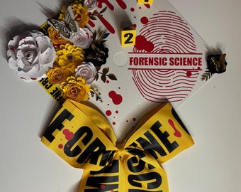 Puede incluir: Un gorro de graduación decorado con flores de papel, una mariposa y una cinta amarilla con el texto "CRIME SCENE". La gorra presenta un diseño de huella dactilar, salpicaduras de sangre y las palabras "FORENSIC SCIENCE".