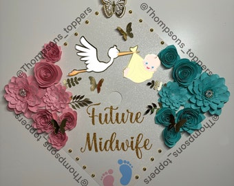Pode incluir: Um chapéu de formatura decorado com flores de papel rosa e turquesa, borboletas douradas e uma cegonha carregando um bebê. O chapéu também apresenta o texto "Future Midwife" em glitter dourado e um par de pegadas de bebê.