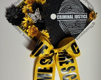 Pode incluir: Um chapéu de formatura preto decorado com flores de papel amarelas e pretas, uma borboleta e uma fita amarela com a inscrição "CRIME SCENE". O chapéu apresenta um design de impressão digital e as palavras "CRIMINAL JUSTICE".