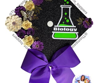 Pode incluir: Um chapéu de formatura preto brilhante decorado com flores de papel creme, roxo e dourado. Apresenta uma borboleta e a palavra "Biology" em verde e branco. Uma grande fita roxa está presa.
