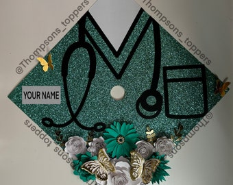 Pode incluir: Um chapéu de formatura com glitter verde-azulado decorado com um estetoscópio preto, uma etiqueta de nome branca e flores de papel. Borboletas douradas e o texto "YOUR NAME" também são apresentados. O chapéu é adornado com flores de papel brancas, verde-azuis e cinzentas.