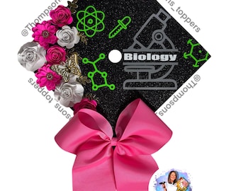 Pode incluir: Um chapéu de formatura preto decorado com gráficos de biologia em verde e prata, incluindo um microscópio e uma cadeia de ADN. O chapéu é adornado com flores de papel rosa e branco, uma borboleta dourada e um grande laço rosa.