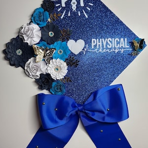 May include: A blue glitter graduation cap decorated with paper flowers in shades of blue, white, and gold. A large blue bow with gold studs is attached. The cap has the words "Physical Therapy" and a caduceus symbol.