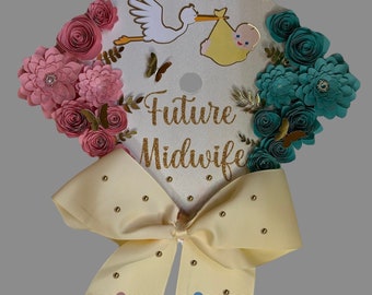 Pode incluir: Um chapéu de formatura decorado com flores de papel rosa e verde-azulado, borboletas douradas e uma cegonha carregando um bebê. O texto "Future Midwife" está em glitter dourado. Um laço creme com pegadas de bebê completa o design.