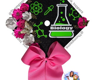 Pode incluir: Um chapéu de formatura preto decorado com um laço rosa, flores rosa e brancas e gráficos com tema de biologia em verde. A palavra "Biology" é exibida em um béquer verde.