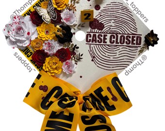 Pode incluir: Um chapéu de formatura decorado com um tema de cena de crime. Apresenta uma fita amarela com o texto "CRIME SCENE", flores de papel, uma borboleta e um desenho de impressão digital com o texto "CASE CLOSED". Inclui marcadores de evidências em miniatura.