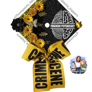 May include: Black graduation cap decorated with yellow and black paper flowers, a butterfly, and gold accents. The cap features the text "FORENSIC PSYCHOLOGY" and "CRIME SCENE" ribbon. The cap is decorated with glitter.