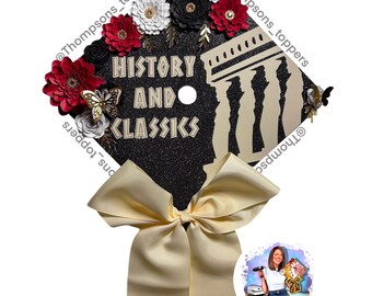 Pode incluir: Um chapéu de formatura preto decorado com flores de papel vermelho, branco e preto, glitter dourado e uma silhueta de um prédio. O chapéu tem uma fita dourada e o texto "HISTORY AND CLASSICS".