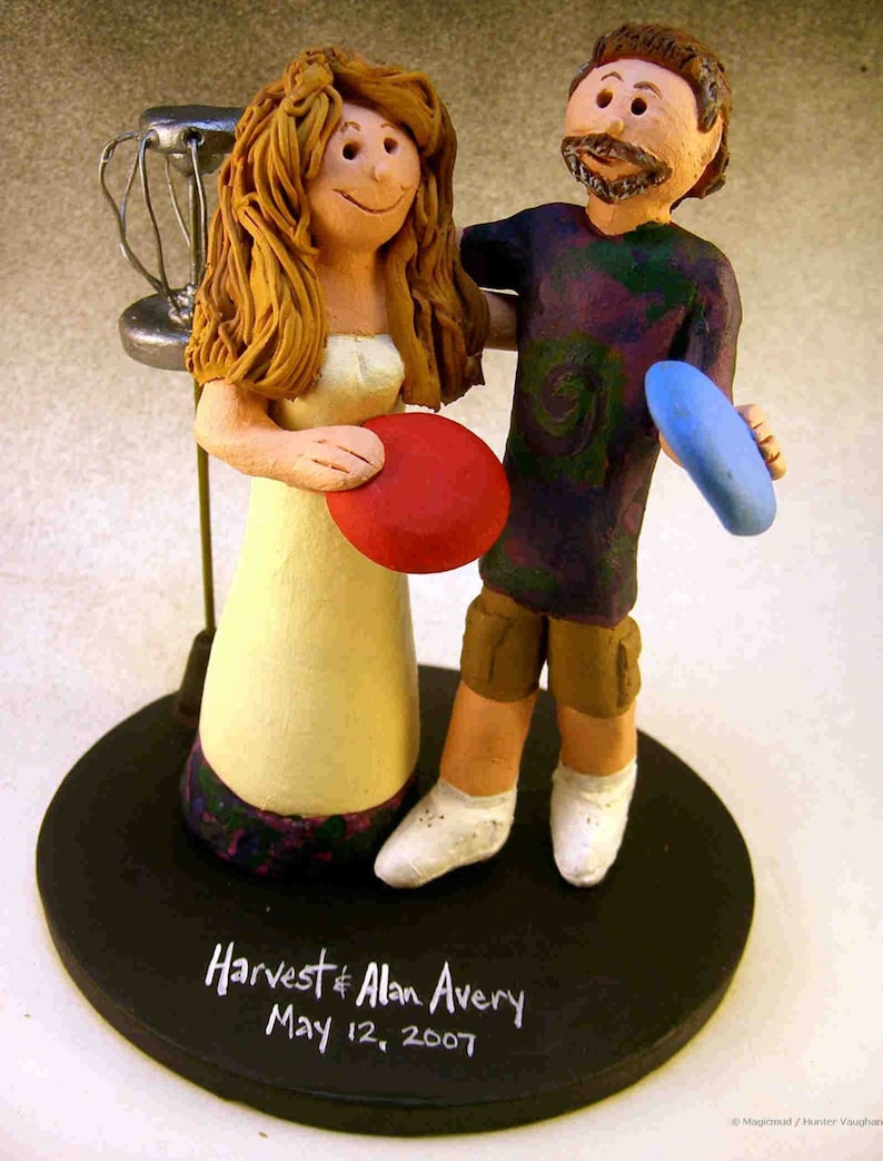 May include: A wedding cake topper featuring a bride and groom dressed in casual attire. The bride is holding a red frisbee and the groom is holding a blue frisbee. The cake topper is on a black base with the text "Harvest & Alan Avery May 12, 2007".