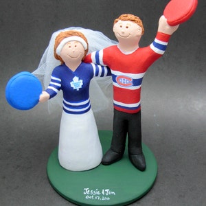 May include: A wedding cake topper featuring a bride and groom dressed in hockey jerseys. The bride is wearing a white dress and a blue and white Toronto Maple Leafs jersey. The groom is wearing a red and white Montreal Canadiens jersey. The bride is holding a blue hockey puck and the groom is holding a red hockey puck. The cake topper is on a green base with the text "Jessie & Jim Oct. 17, 2010" and "www.magicmud.com &copy; Hunter Vaughan".