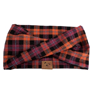 May include: A plaid headband in shades of orange, red, and black. The fabric features a classic tartan pattern with thin blue lines. A small brown leather tag with a bear logo is sewn on the front.