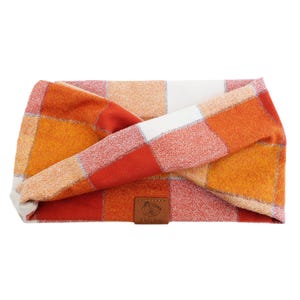 May include: A plaid headband in shades of orange, red, and white. The fabric is a soft, woven material with a rectangular pattern. A small brown leather tag with text is sewn on the headband.