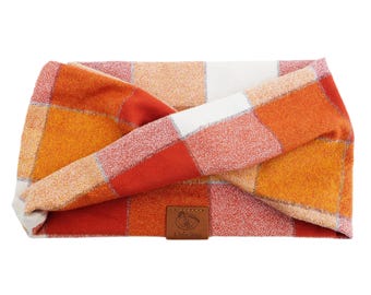 May include: A twisted headband featuring a plaid pattern in shades of orange, red, and white. The fabric has a textured appearance and includes a small, rectangular brown leather tag at the bottom centre with the text A Lucky Dog.
