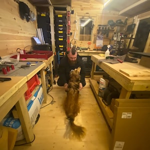 May include: A man with a mohawk hairstyle is kneeling on the floor of a workshop, petting a golden-brown dog. The workshop is filled with tools and equipment, including a workbench with a table saw, a drill press, and a variety of other tools. The dog is looking up at the man with a happy expression.