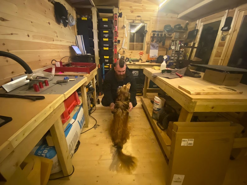 May include: A man with a mohawk hairstyle is kneeling on the floor of a workshop, petting a golden-brown dog. The workshop is filled with tools and equipment, including a workbench with a table saw, a drill press, and a variety of other tools. The dog is looking up at the man with a happy expression.
