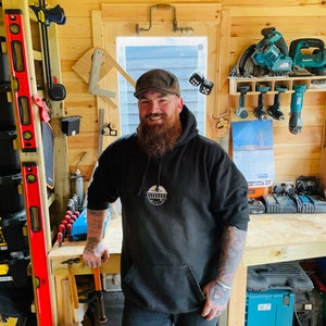May include: A man with a beard wearing a black hoodie with a white logo that says "Woodworking" in a circle. He is standing in a woodshop with tools on the walls and a workbench in front of him.