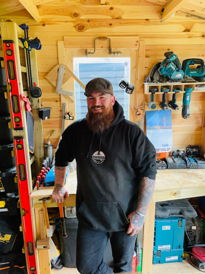 May include: A man with a beard wearing a black hoodie with a white logo that says "Woodworking" in a circle. He is standing in a woodshop with tools on the walls and a workbench in front of him.