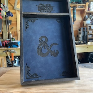 May include: A wooden dice tray with a blue felt lining and a black leatherette inlay featuring the Dungeons & Dragons logo and a stylized dragon and ampersand.