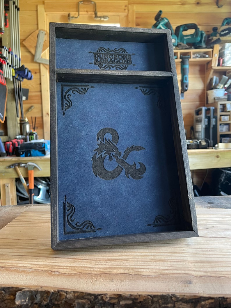 May include: A wooden dice tray with a blue felt lining and a black leatherette inlay featuring the Dungeons & Dragons logo and a stylized dragon and ampersand.