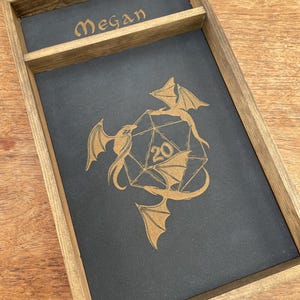 May include: A wooden dice tray with a black felt interior. The tray has a small compartment for dice and a larger compartment for rolling dice. The tray is engraved with the name "Megan" and a design of two dragons surrounding a twenty-sided die.