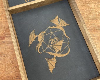 May include: A wooden dice tray with a black felt interior. The tray has a small compartment for dice and a larger compartment for rolling dice. The tray is engraved with the name "Megan" and a design of two dragons surrounding a twenty-sided die.