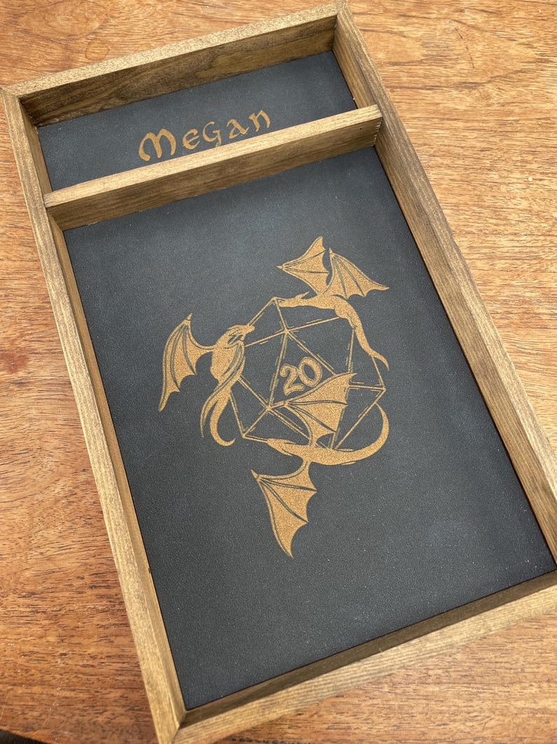 May include: A wooden dice tray with a black felt interior. The tray has a small compartment for dice and a larger compartment for rolling dice. The tray is engraved with the name "Megan" and a design of two dragons surrounding a twenty-sided die.