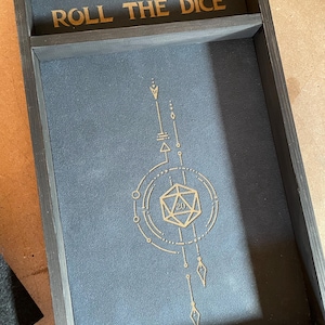 May include: A wooden dice tray with a black felt lining and a gold design featuring a 20-sided die and the text "ROLL THE DICE".