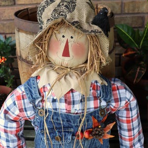 May include: A scarecrow doll wearing a burlap hat with a black pattern, a red, white, and blue plaid shirt, and blue denim overalls. The doll has a fabric face with a red triangular nose, straw hair, and a small black bird attached to the hat.