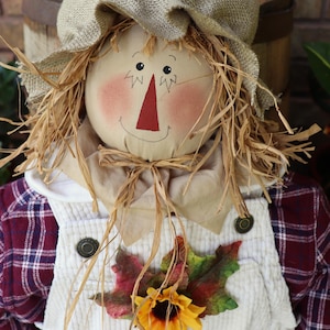 May include: A scarecrow doll wearing a tan burlap hat, a white corduroy overall, and a maroon and white plaid shirt. The doll has a round fabric face with a red triangular nose, painted eyes, and straw hair. A sunflower and autumn leaves are attached.