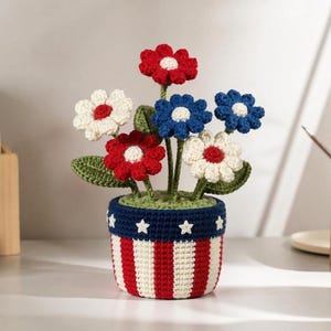 Könnte beinhalten: Gehäkelte Blumenanordnung in patriotischen Farben. Die Blumen sind rot, weiß und blau, mit grünen Blättern, in einem Topf mit rot-weißen Streifen und blauen Sternen. Handgefertigte Handwerkskunst.