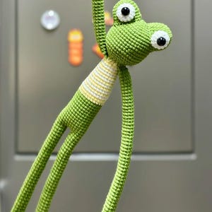 May include: A handmade green crochet frog with large white and black eyes. The frog has long, thin legs and arms, and a striped yellow and white section at the neck. The frog is hanging against a neutral background.