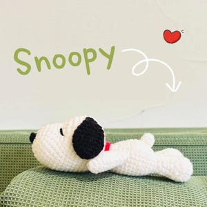 May include: A crocheted Snoopy toy in white and black, with a red collar, resting on a green textured surface. The word "Snoopy" is written in green above the toy, with a red heart and arrow graphic.