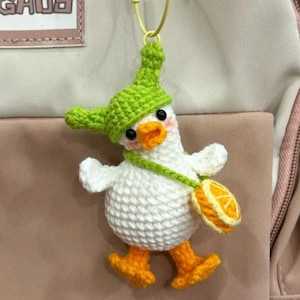 Può includere: Un portachiavi a forma di anatra all'uncinetto fatto a mano, con un cappello verde e zampe arancioni, che porta una piccola borsa arancione. L'anatra è bianca con un becco arancione e occhi neri. Il portachiavi è attaccato a uno zaino con la scritta "MENGHUO".